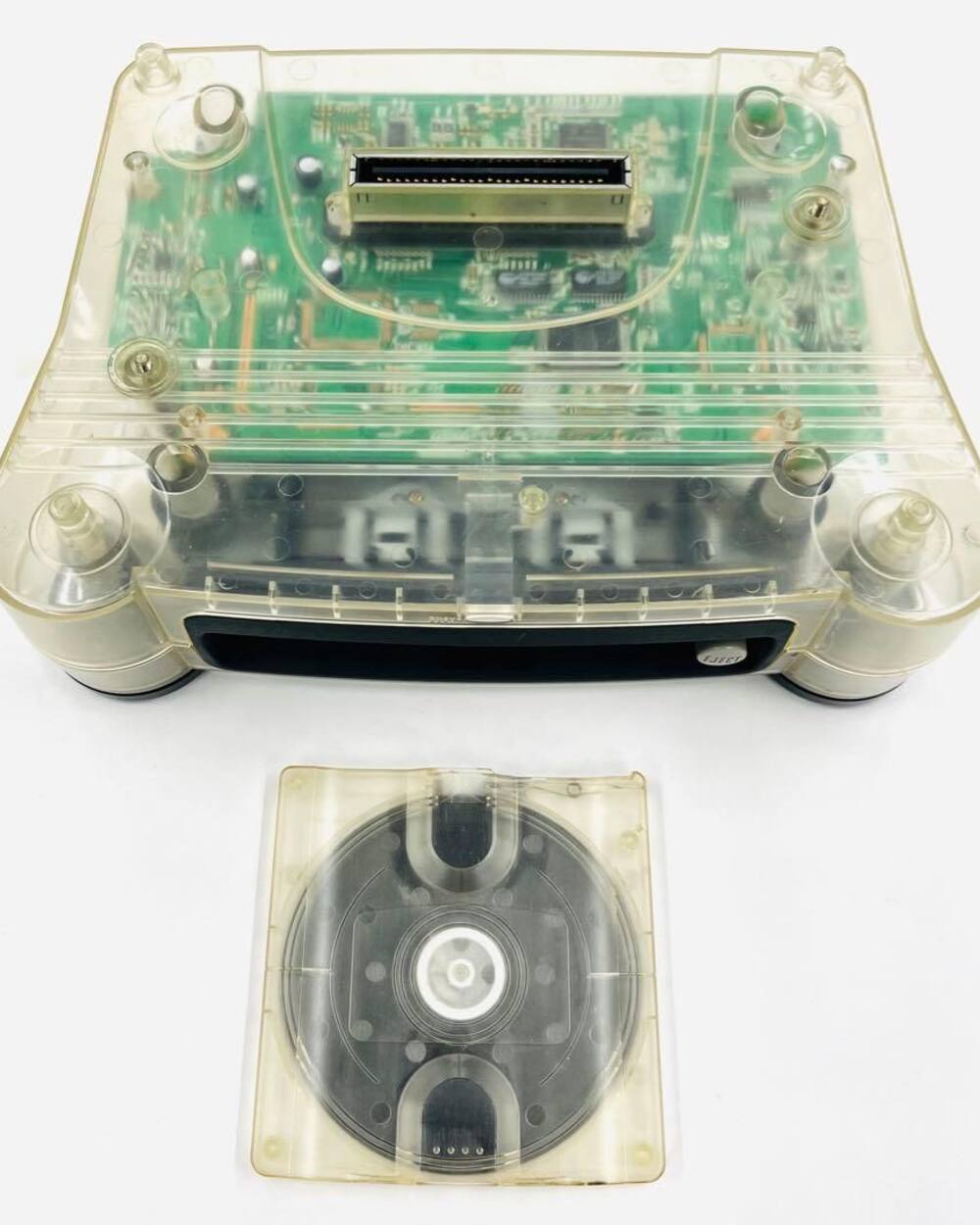 A transparent Nintendo 64DD disk drive seen from above, its green circuit board and expansion connector visible through the clear shell, with a clear blank disc lying below it