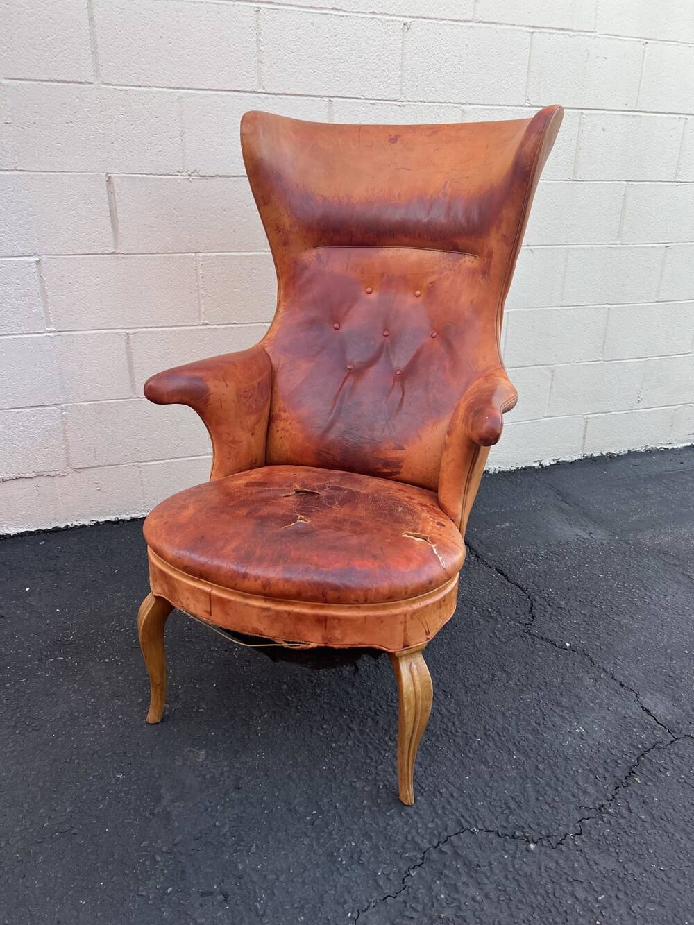 The Henningsen wing chair as Justin Miller collected it, standing on asphalt against a white block wall, the leather worn and split across the seat